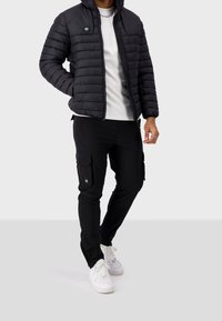 Giubbotto invernale nero con zipper e cappuccio, caratterizzato da un motivo testurizzato e un badge. Indossato sopra una maglietta bianca e pantaloni cargo neri.