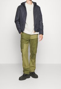 Mann in navyfarbener Kapuzenjacke, beigem T-Shirt, olivgrünen Patchwork-Cargo-Hosen und schwarzen klobigen Stiefeln, der vor einem schlichten hellen Hintergrund steht.