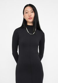 Czarna długa sukienka z długim rękawem i wysokim dekoltem, dopasowany krój oraz gładka tekstura. Uzupełniona naszyjnikiem z pereł i srebrnego koloru.