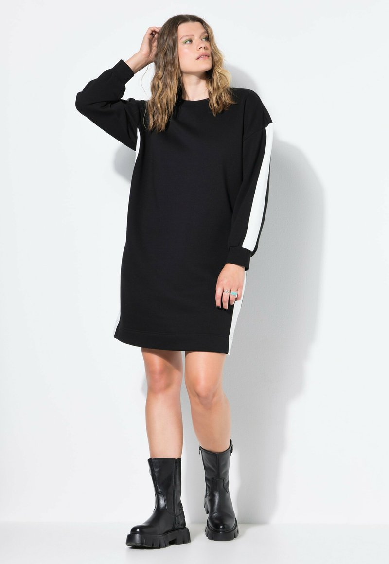 Vestido sudadera negro con mangas largas, con rayas blancas en los laterales. Tejido suave, corte holgado, longitud hasta medio muslo. Llevado con botas negras hasta el tobillo.