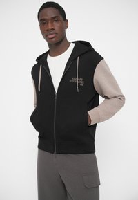 Sudadera con capucha negra con cremallera y mangas beige, con bordado de logo. Hecha de tela suave con bolsillo tipo canguro y capucha ajustable con cordón.
