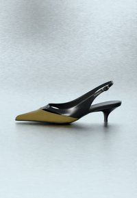 Czółenka na niskim obcasie typu kitten heel z odkrytą piętą i szpiczastym noskiem, z oliwkowozieloną przednią częścią i czarną cholewką na jasnoszarym tle.