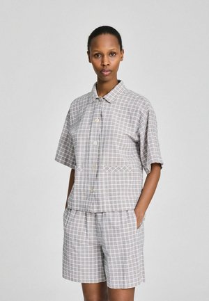 Kvinde med kort hår iført grå ternet kortærmet skjorte og matchende shorts, står med hænderne i lommerne mod en ensfarvet baggrund.