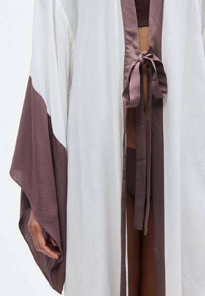 Ροή κάπα σε στυλ κιμονό σε λευκό και καφέ με φαρδιά μανίκια, δεμένη μπροστά με κορδέλα σε ταιριαστή καφέ απόχρωση. Υφή υφάσματος με σχεδιασμό.