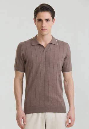 Młody mężczyzna w brązowej, krótkorękawowej, dzianej koszulce polo z pionowymi paskami, stojący na tle jasnego, jednolitego tła.