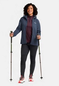Giacca softshell blu scuro con cappuccio, strato intermedio color bordeaux, pantaloni neri e scarpe da ginnastica rosse e bianche; tiene in mano i bastoncini da trekking.