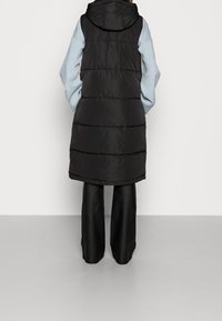 Pikowana czarna kamizelka z kapturem, o warstwowej konstrukcji i przeszywanym wzorze, zestawiona z jasnoniebieskimi długimi rękawami oraz czarnymi szerokimi spodniami.