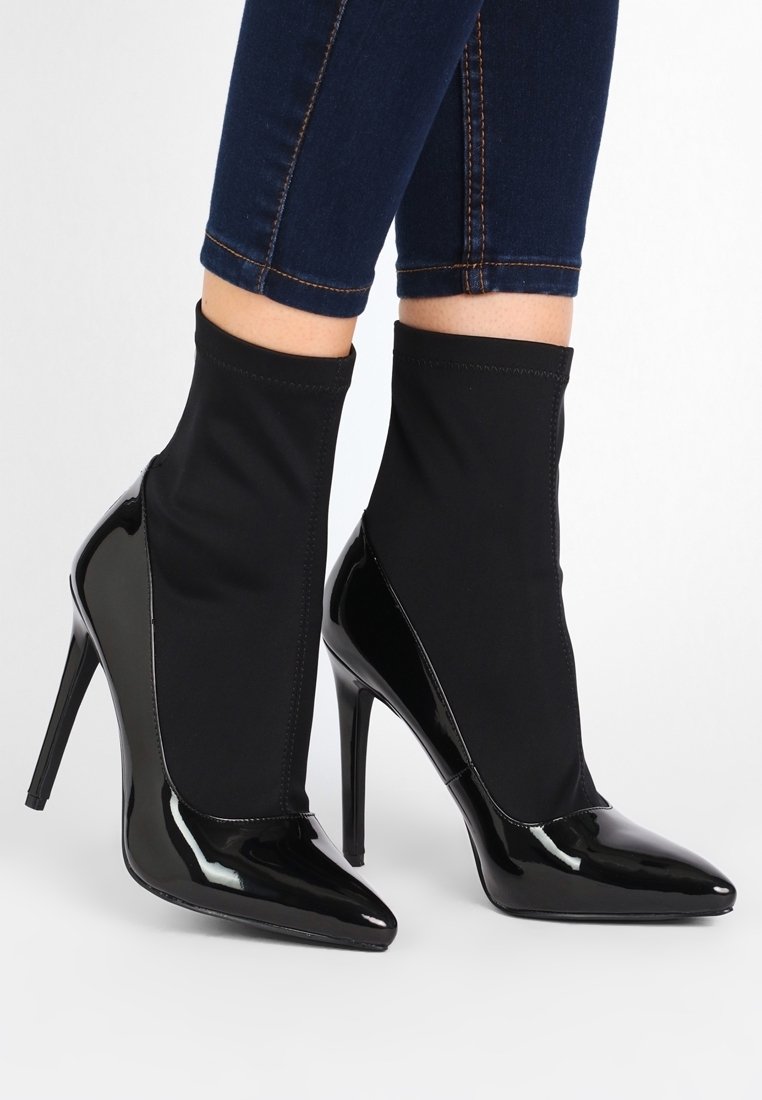 Bottines à talons hauts en cuir verni noir avec tiges en tissu extensible, portées avec un jean skinny bleu foncé.