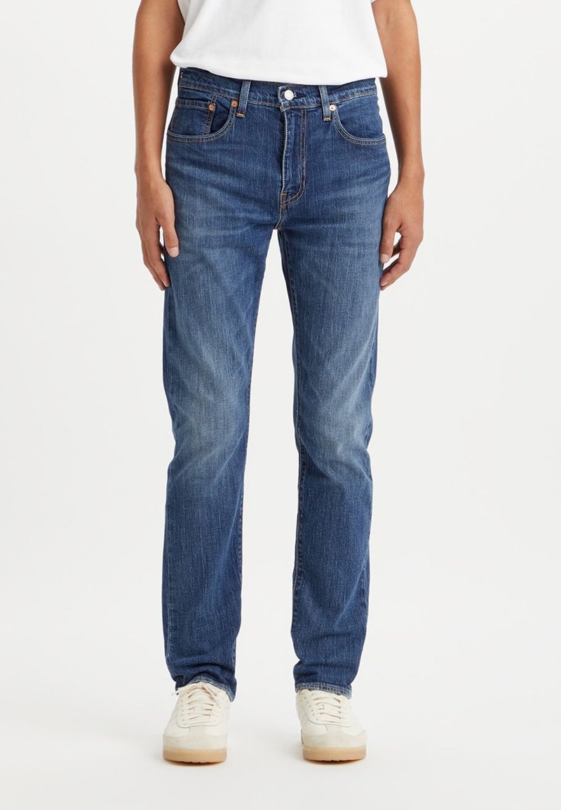 Jeans de mezclilla azul oscuro con un diseño de pierna recta, estilo de cinco bolsillos y un ligero desgaste en el frente. Combinados con zapatillas blancas.
