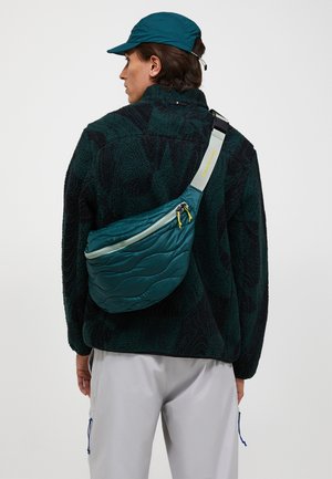 Persona que lleva una chaqueta de forro polar verde oscuro con estampado, bolso bandolera acolchado de color azul verdoso, gorra azul verdoso y pantalones gris claro, de espaldas sobre un fondo blanco.