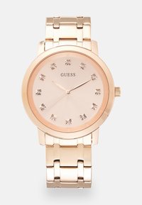 Montre en or rose avec un bracelet métallique, un cadran rose clair texturé et des marqueurs en cristal à chaque heure, affichant un design minimaliste.