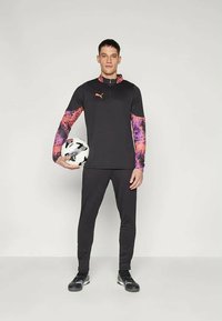 Μαύρη αθλητική στολή με πολύχρωμα μοτίβα στα μανίκια, που περιλαμβάνει φούτερ με φερμουάρ και εφαρμοστά παντελόνια. Κρατώντας μια μπάλα ποδοσφαίρου.