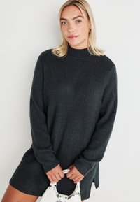 Dark gray knitted sweater dress with a high neckline, long sleeves, side slits, and ribbed texture, held with a white handbag.