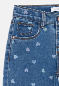 Jeans aus Denim in Blau mit einem subtilen Herzmuster in Pastellfarben. Verfügt über einen klassischen Schnitt, fünf Taschen und kontrastierenden orangen Nähten.