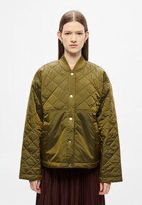 Puffasztott olívazöld dzseki patentos záródással, bordázott gallérral és széles ujjal, barna pliszírozott szoknyával párosítva.