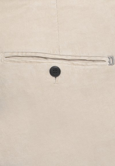 Tessuto di cotone beige con texture rigata. Presenta una chiusura con bottone e una piccola tasca con cuciture visibili. Accento argentato vicino alla tasca.