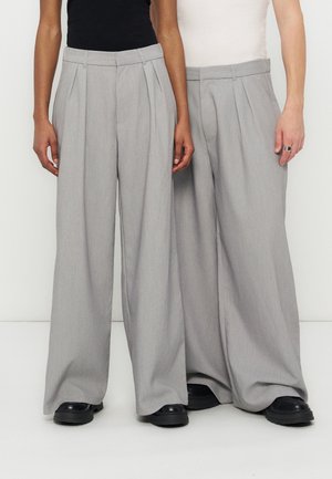 Δύο άτομα φορούν ταιριαστά γκρι παντελόνια με φαρδιά μπατζάκια και μαύρα παπούτσια, στέκονται δίπλα δίπλα σε απλό φόντο.