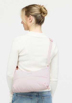 Frau mit blondem Haar, zu einem Dutt gebunden, trägt einen weißen, gerippten Pullover und eine hellrosa gesteppte Umhängetasche, gesehen von hinten.