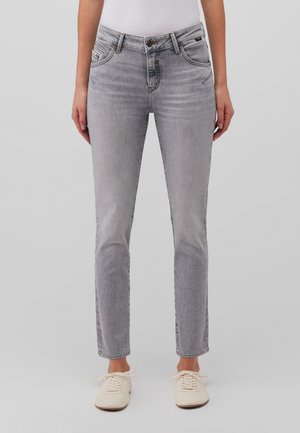 Vue de face d'une personne portant un jean slim gris clair et des baskets blanches sur un fond gris uni.