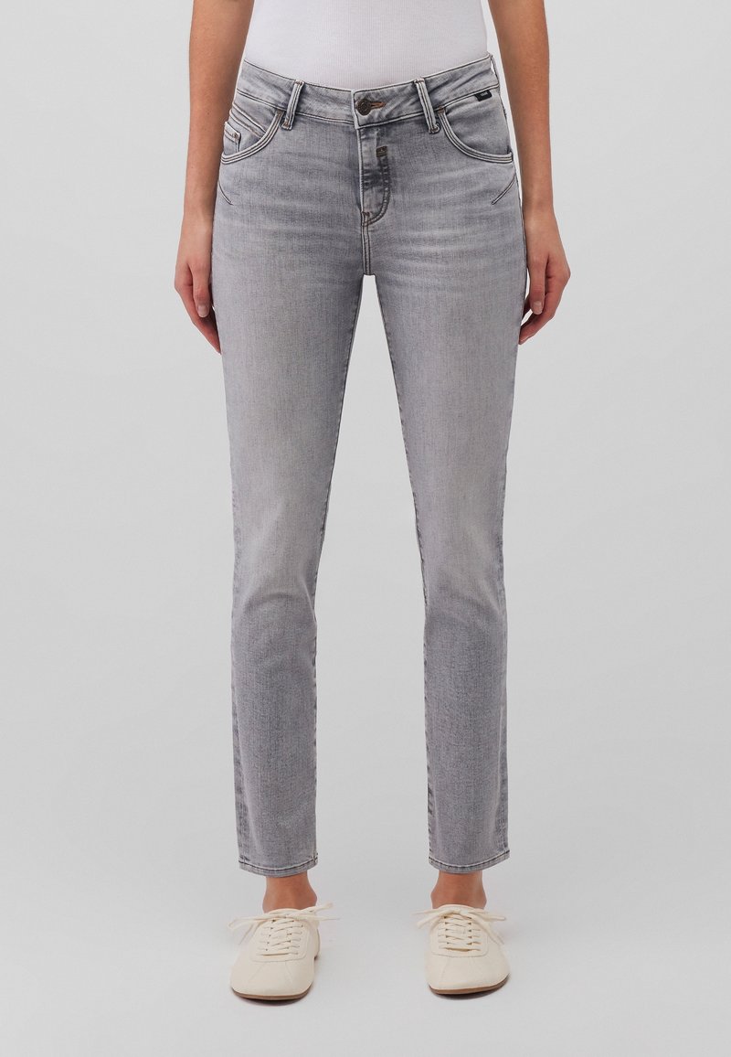 Vue de face d'une personne portant un jean slim gris clair et des baskets blanches sur un fond gris uni.