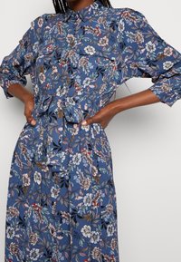 Šaty s květinovým potiskem z modré tkaniny, s límečkem na knoflíky, dlouhými rukávy a detailem na zavázání v pase. Obsahují barevné květinové vzory.