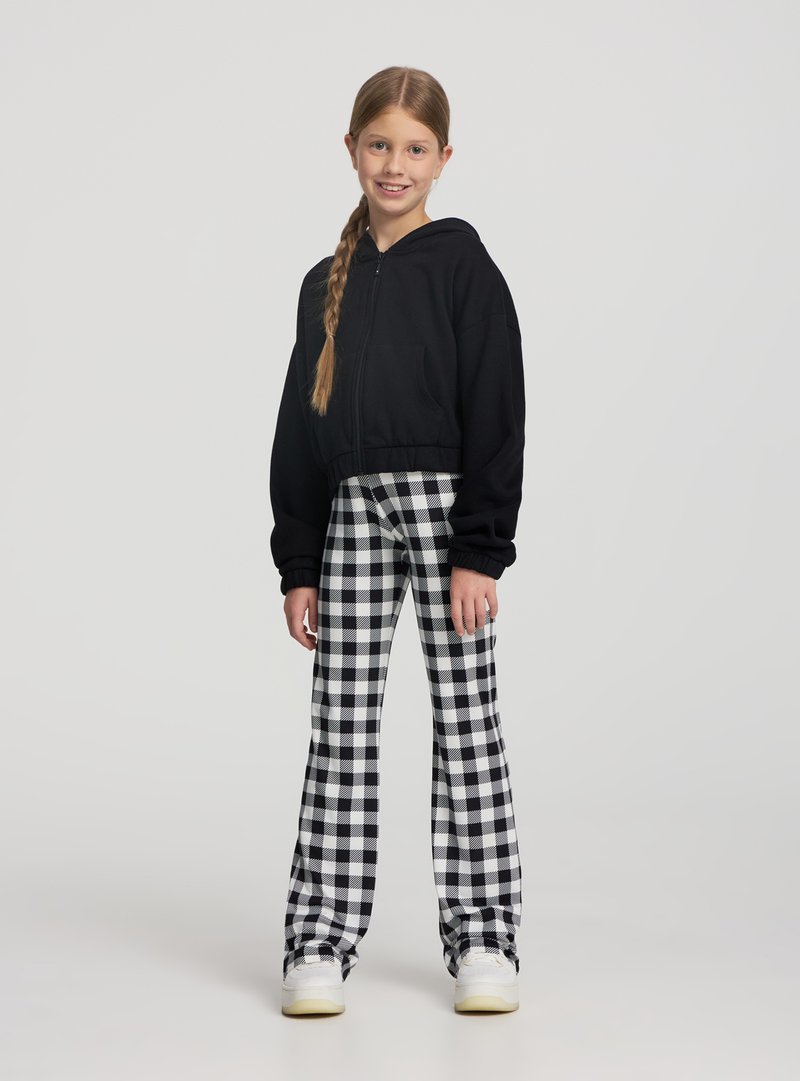 Felpa nera corta con zip, abbinata a pantaloni a vita alta a quadretti bianchi e neri. Scarpe da ginnastica bianche completano il look.