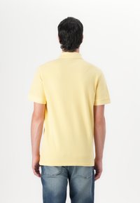 Gul polo-skjorte med en myk tekstur, korte ermer og en klassisk krage, kombinert med blå jeans. Bakfra vises enkle design detaljer.