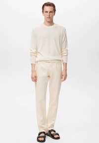 Beżowy sweter i dopasowane spodnie, gładka tekstura, slim fit, noszone z czarnymi sandałami. Prosty i monochromatyczny design na neutralnym tle.