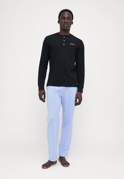 Camisa henley de manga longa preta com três botões e "POLO" em amarelo, combinada com calças de algodão azul claro, em pé descalço contra um fundo simples.