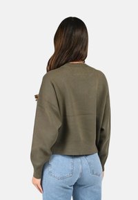 Pull court vert olive avec un col rond et des manches longues et larges. Texture lisse avec un ourlet côtelé. Associé à un jean bleu clair.