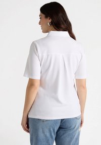 Camicia bianca a maniche corte con colletto, in tessuto morbido, caratterizzata da una piega sul retro e un orlo curvo, abbinata a jeans azzurri chiari.