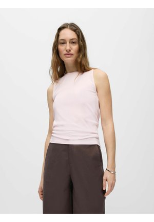 Femme aux cheveux longs et ondulés portant un top rose pâle sans manches et un pantalon large foncé, sur un fond clair uni.