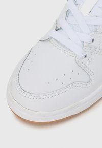 Sneaker en cuir blanc avec une boîte à orteils texturée, des détails perforés et des lacets blancs plats. Il présente une semelle en caoutchouc tan et des coutures contrastées.