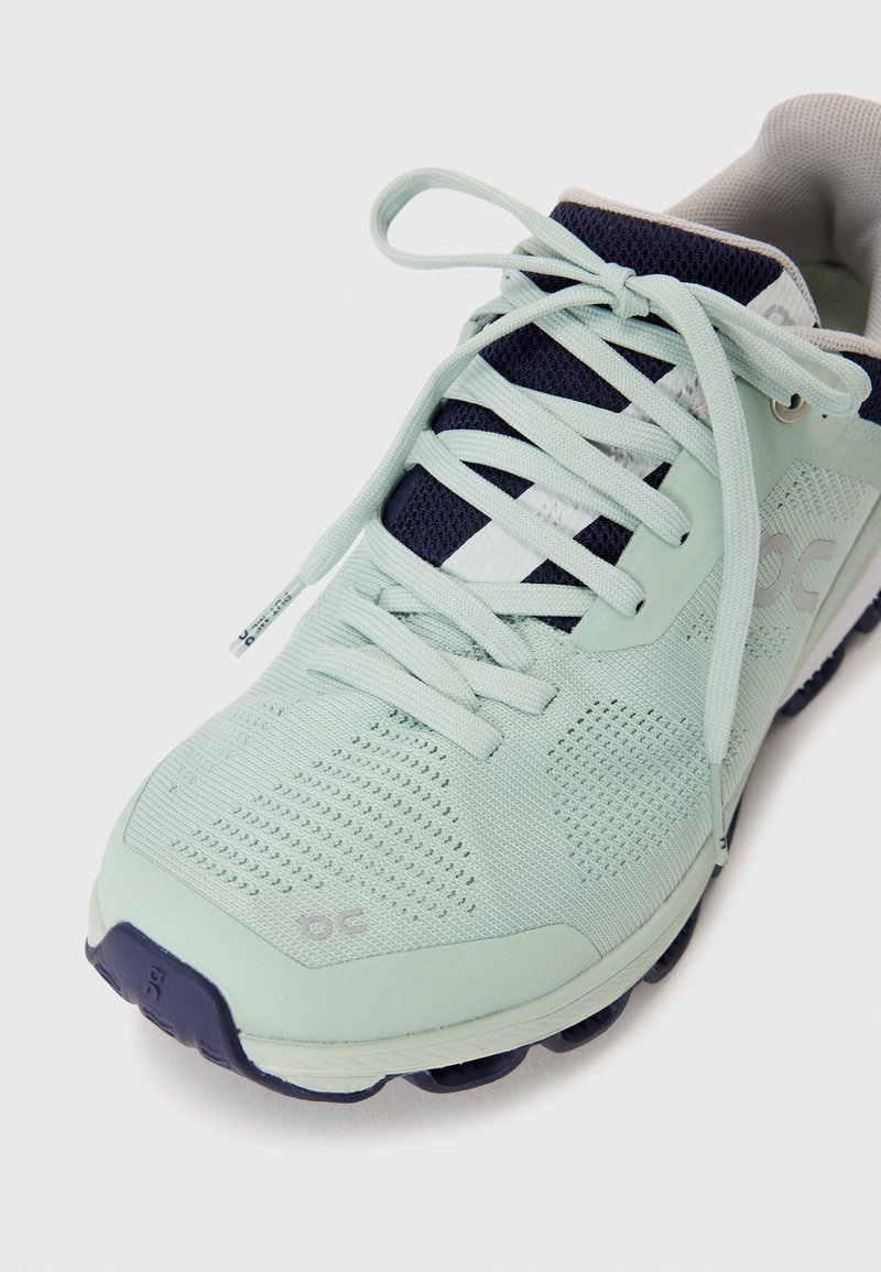 Hellgrüner Sportschuh mit atmungsaktivem Mesh-Obermaterial, marineblauen Akzenten, flachen Schnürsenkeln und einer strukturierten Sohle für besseren Halt. Merkmale ein Logo-Detail an der Seite.