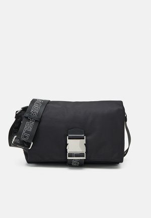 Czarna prostokątna torba typu crossbody z srebrną klamrą i regulowanym paskiem z nadrukiem logo na jednolitym białym tle.