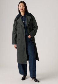Donkergroene trenchcoat met dubbelzijdige knopen, brede kraag en zijzakken, gecombineerd met blauwe spijkerbroeken en zwarte haklaarzen.
