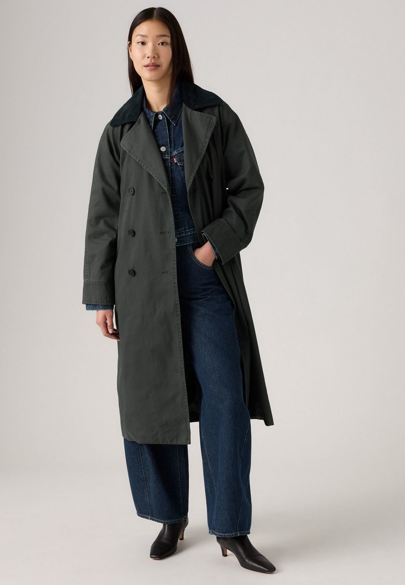 Manteau trench vert foncé avec des boutons double boutonnage, un large col et des poches latérales, associé à des salopettes en denim bleu et des bottes à talons noires.