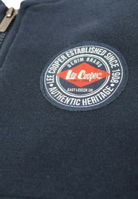 Gros plan sur un tissu bleu foncé avec un patch circulaire du logo Lee Cooper indiquant « Établi depuis 1908 » et « Patrimoine Authentique ».