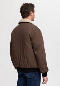Hombre con chaqueta marrón con cuello de borrego beige y vaqueros azules, de pie de espaldas sobre un fondo liso.