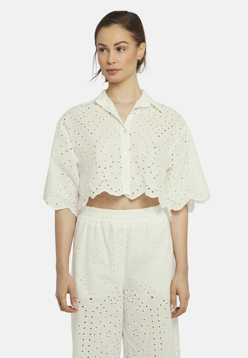 Witte cropped shirt met gegolfde zoom, voorzien van oogjes borduursel en een knoopsluiting aan de voorkant. In combinatie met bijpassende loszittende broek.