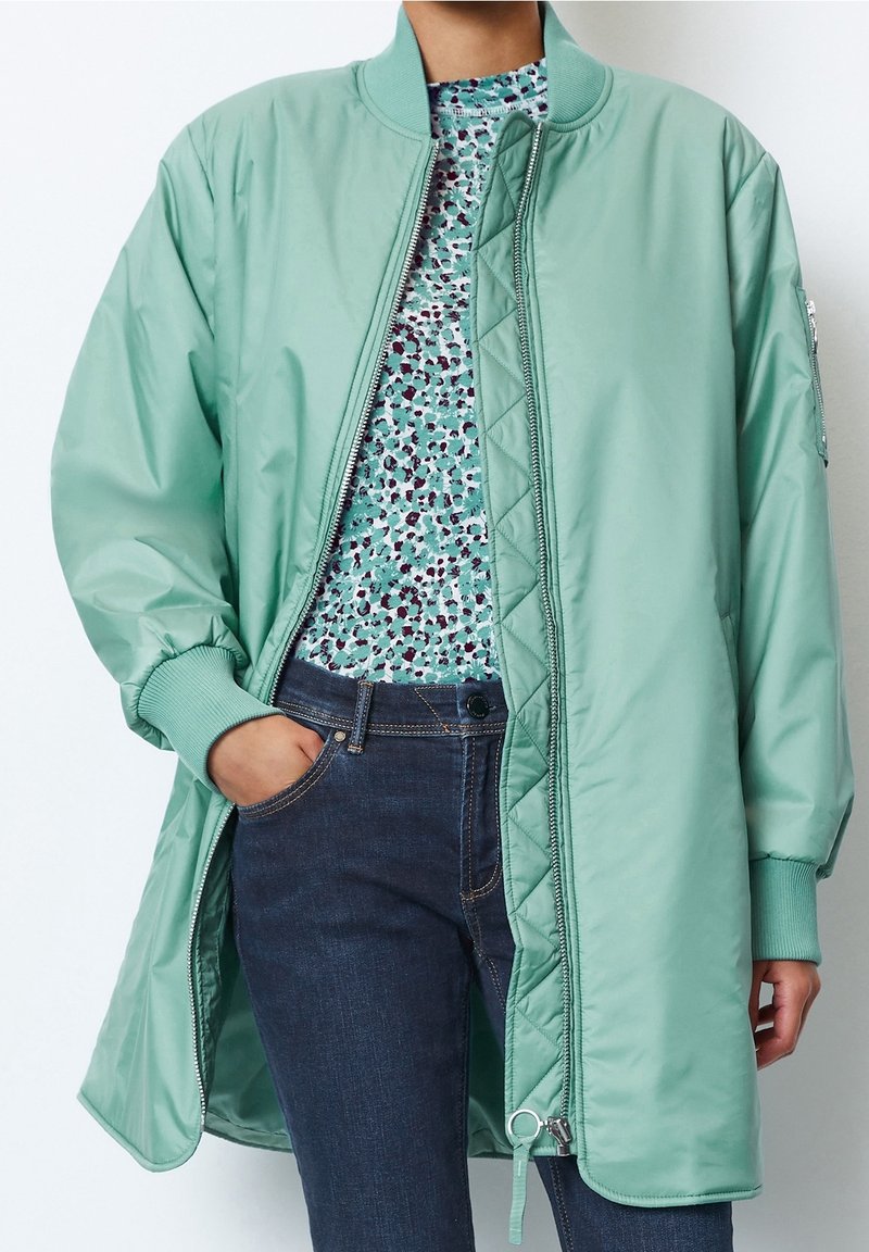 Bomber bunda v mátově zelené barvě z lesklé látky, s uzavíráním na zip, žebrovanými manžetami a prošívanou vnitřní podšívkou, navršená na vzorovaném topu.