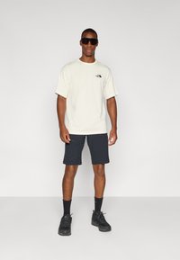 Light beige t-shirt with a small black logo, paired with dark navy shorts. Model wears black sunglasses and black sneakers with dark socks.