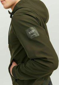 Green softshell hoodie with a full zipper, drawstring hood, side pockets, and a patch sleeve label featuring coordinates.