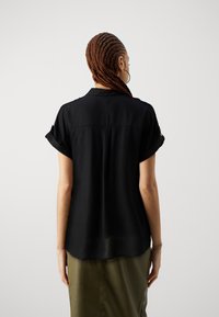 Camisa negra de manga corta con un corte relajado y puños enrollados, hecha de una tela suave; cuenta con una parte posterior con botones y un brillo sutil.