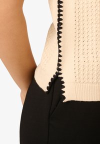 Beige gestricktes ärmelloses Oberteil mit schwarzem gewelltem Rand, getragen mit schwarzen Hosen, zeigt Nahaufnahme von der Seite und von hinten.