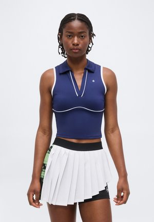 Femme aux cheveux tressés portant un crop top bleu marine sans manches et une jupe de tennis plissée blanche avec un short noir en dessous.
