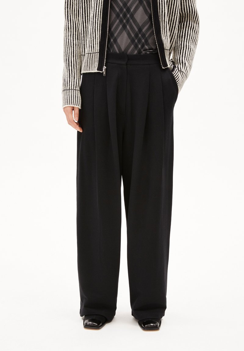 Pantalon noir à jambes larges associé à une chemise à carreaux rentrée et un pull zippé beige et noir à rayures.