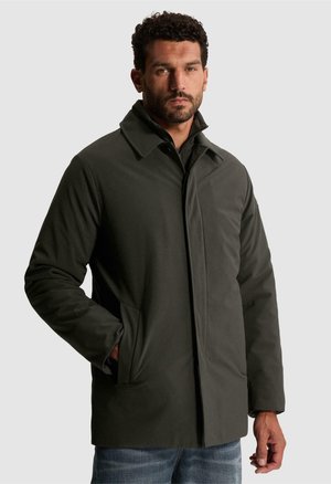 Homme portant un long manteau vert foncé à col montant et les mains dans les poches, debout devant un fond gris clair uni.