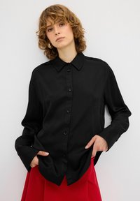 Camicia nera a maniche lunghe con colletto, texture liscia e vestibilità leggermente rilassata, abbinata a una gonna rossa.