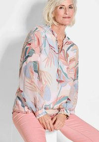 Bluse mit floralen Mustern und Rüschensaum am Hals und an den Ärmeln, in sanften Farben von Rosa, Blau und Grün, kombiniert mit hellrosa engen Hosen.
